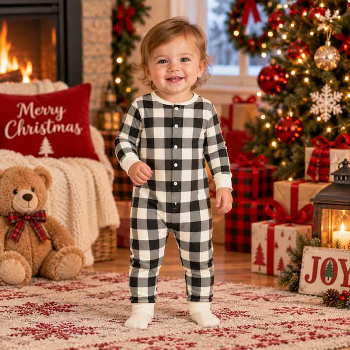 A toddler stands in a cozy Christmas living room wearing a black and white buffalo plaid button-front one-piece pajama. The background features a decorated Christmas tree, wrapped gifts, a teddy bear, and a fireplace with a 'Merry Christmas' pillow.