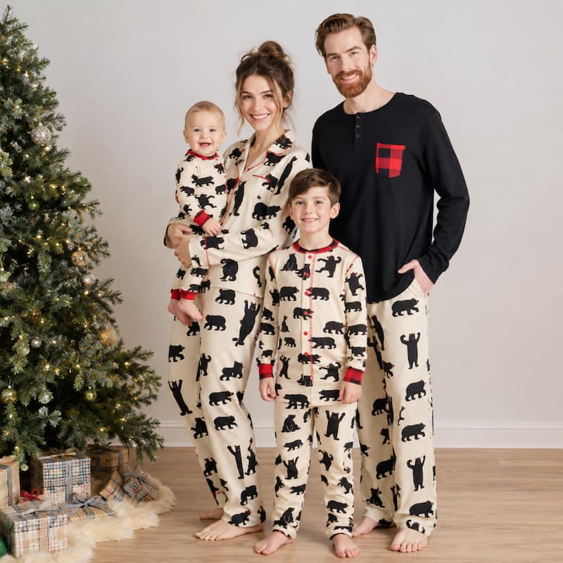 A family of four wearing matching holiday pajamas stands indoors next to a decorated Christmas tree. The pajamas are cream with a black bear print, with red trim and buttons.