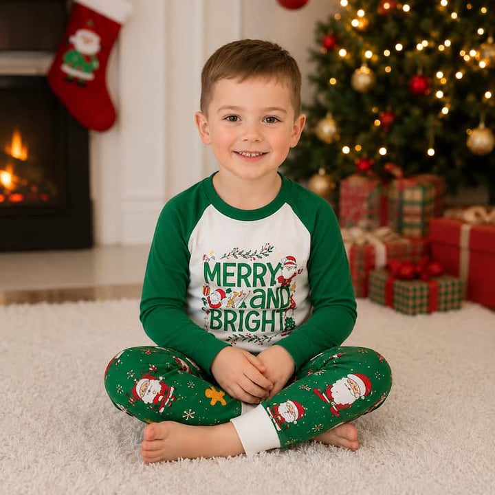 A young boy sits cross-legged on a white carpet in front of a Christmas tree and fireplace, wearing a green and white raglan long-sleeve pajama top with 'MERRY AND BRIGHT' printed on the front, and matching green pajama pants with Santa Claus illustrations.
