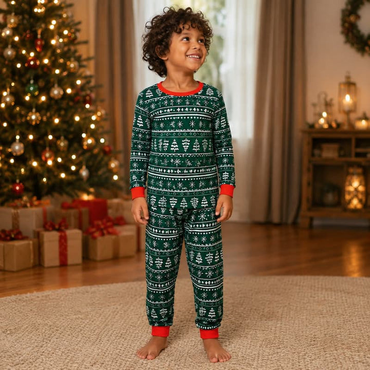 A young child wearing a green and white Christmas tree patterned pajama set stands barefoot on a rug in a cozy holiday room with a decorated Christmas tree and wrapped gifts in the background.