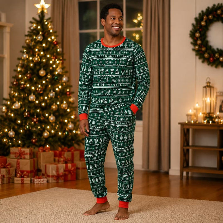 A man wearing a green and white Christmas-themed patterned pajama set stands barefoot in a cozy living room with a decorated Christmas tree, wrapped gifts, and warm lights in the background.