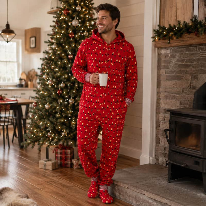 A man wearing a red Christmas-themed hooded pajama set with a white and gold snowman pattern, standing in a cozy living room with a lit Christmas tree and fireplace.
