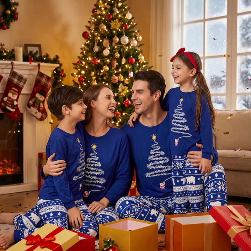 A family of four wearing matching blue Christmas pajamas sits together in a cozy living room in front of a decorated Christmas tree. The set includes long-sleeve tops with white tree and snowflake patterns and matching patterned pants.