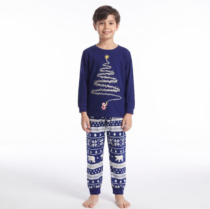 A young boy stands barefoot against a plain white studio background wearing a navy blue long-sleeve pajama top with a white Christmas tree graphic and matching patterned pajama pants with snowflake, polar bear, and reindeer motifs.