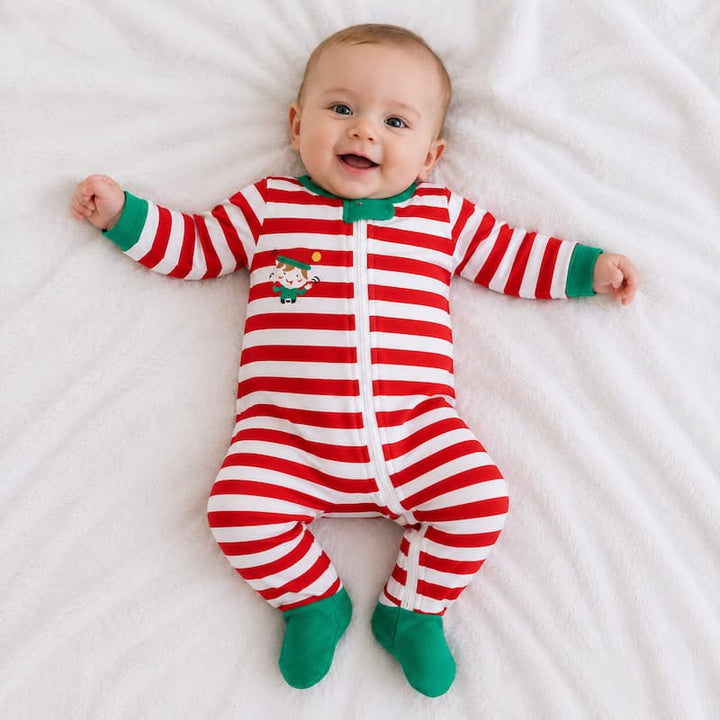 A baby lying on a white blanket wearing a red and white striped footed sleeper with green cuffs and socks, plus a small Santa graphic on the chest.